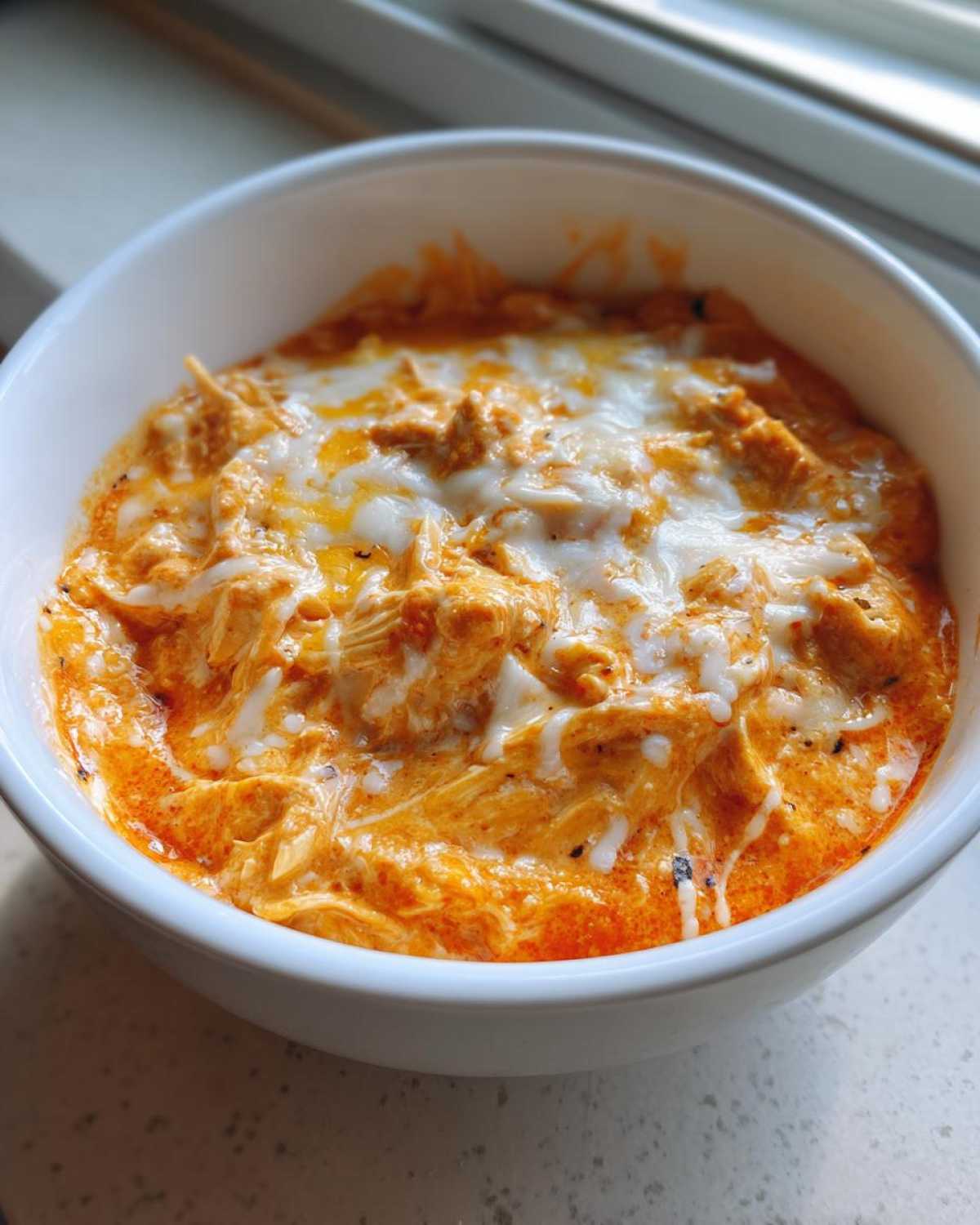 Close-up of creamy, orange buffalo chicken dip topped with melted white cheese in a white bowl.