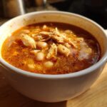 Close-up of a white mug filled with rich, red broth chicken pozole, featuring shredded chicken and hominy.