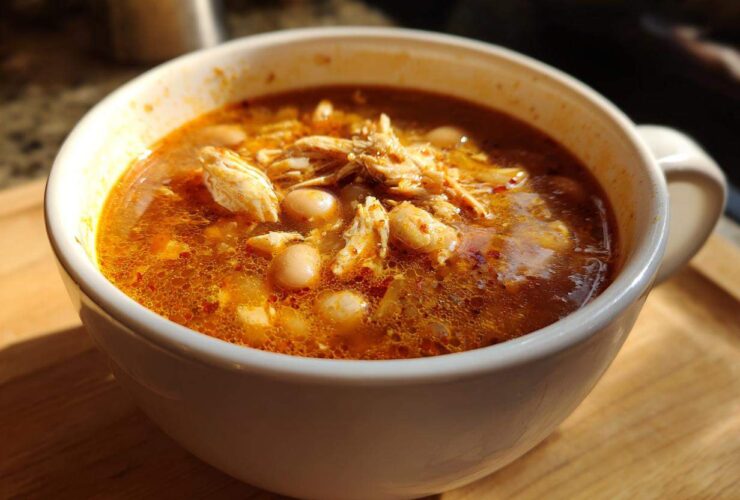 Close-up of a white mug filled with rich, red broth chicken pozole, featuring shredded chicken and hominy.
