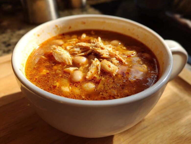 Close-up of a white mug filled with rich, red broth chicken pozole, featuring shredded chicken and hominy.