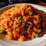 A close-up of a mound of vibrant orange Spanish rice mixed with bright green peas and diced tomatoes on a white plate.