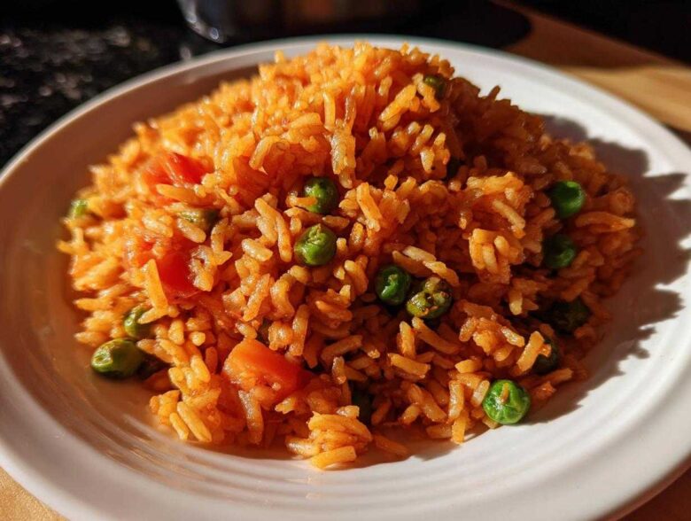 A close-up of a mound of vibrant orange Spanish rice mixed with bright green peas and diced tomatoes on a white plate.