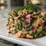 A mound of creamy salmon salad mixed with greens and red onion, topped with fresh dill.