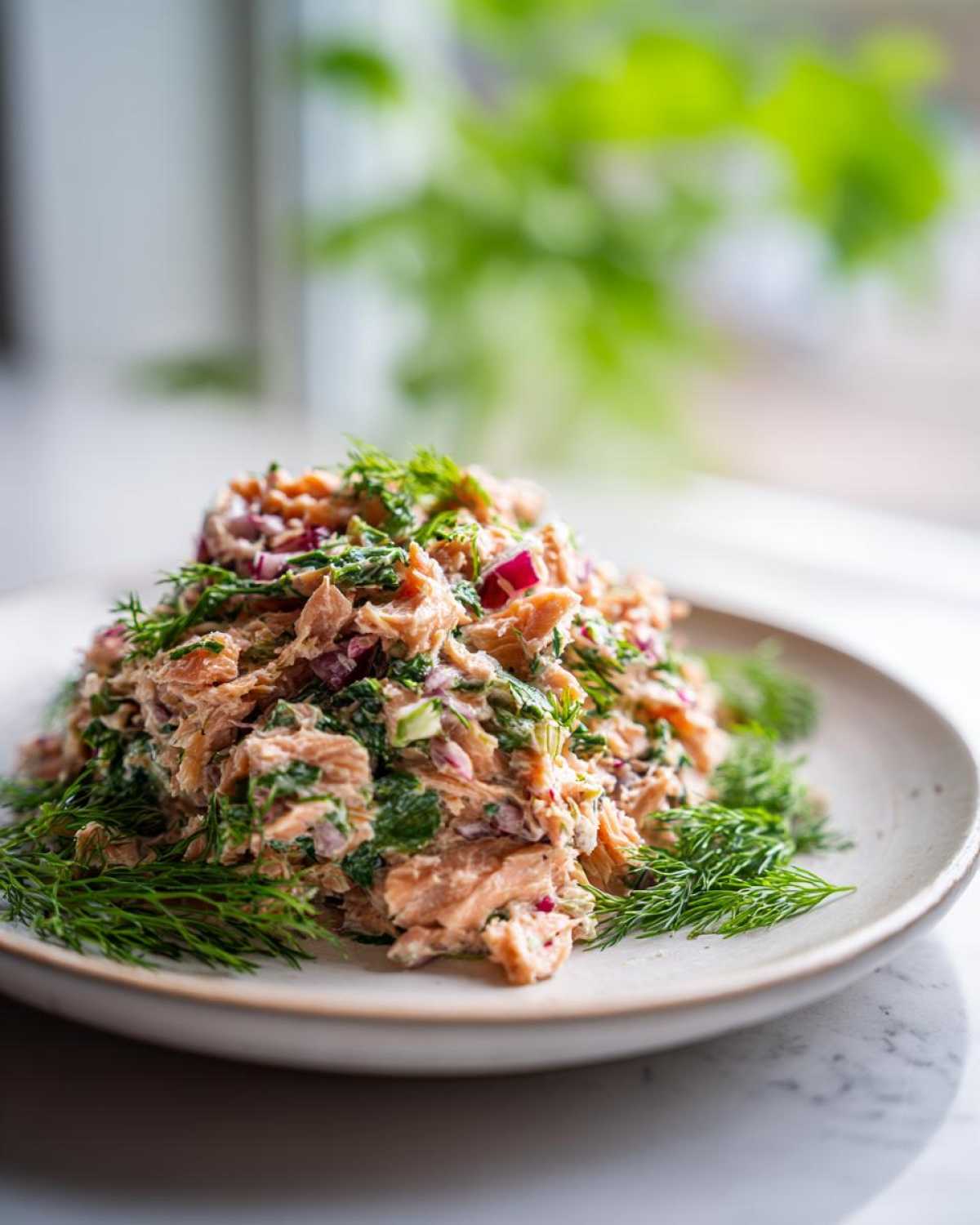 A mound of fresh salmon salad mixed with red onion and herbs, garnished heavily with dill, served on a light plate.