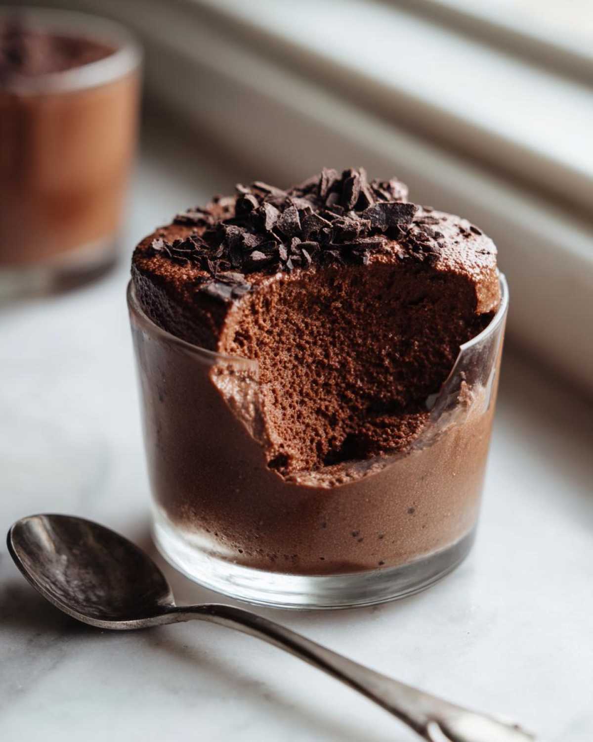 Close-up of rich, airy chocolate mousse in a glass with a bite taken out, topped with chocolate shavings.