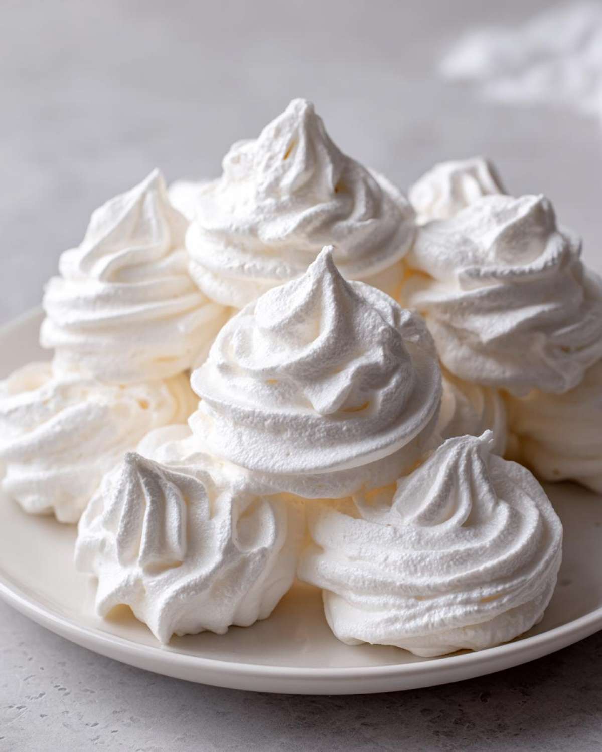 A close-up of several perfectly piped, white meringue cookies stacked on a white plate.