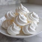 A pile of perfectly piped, bright white meringue cookies with distinct swirls, resting on a white plate.