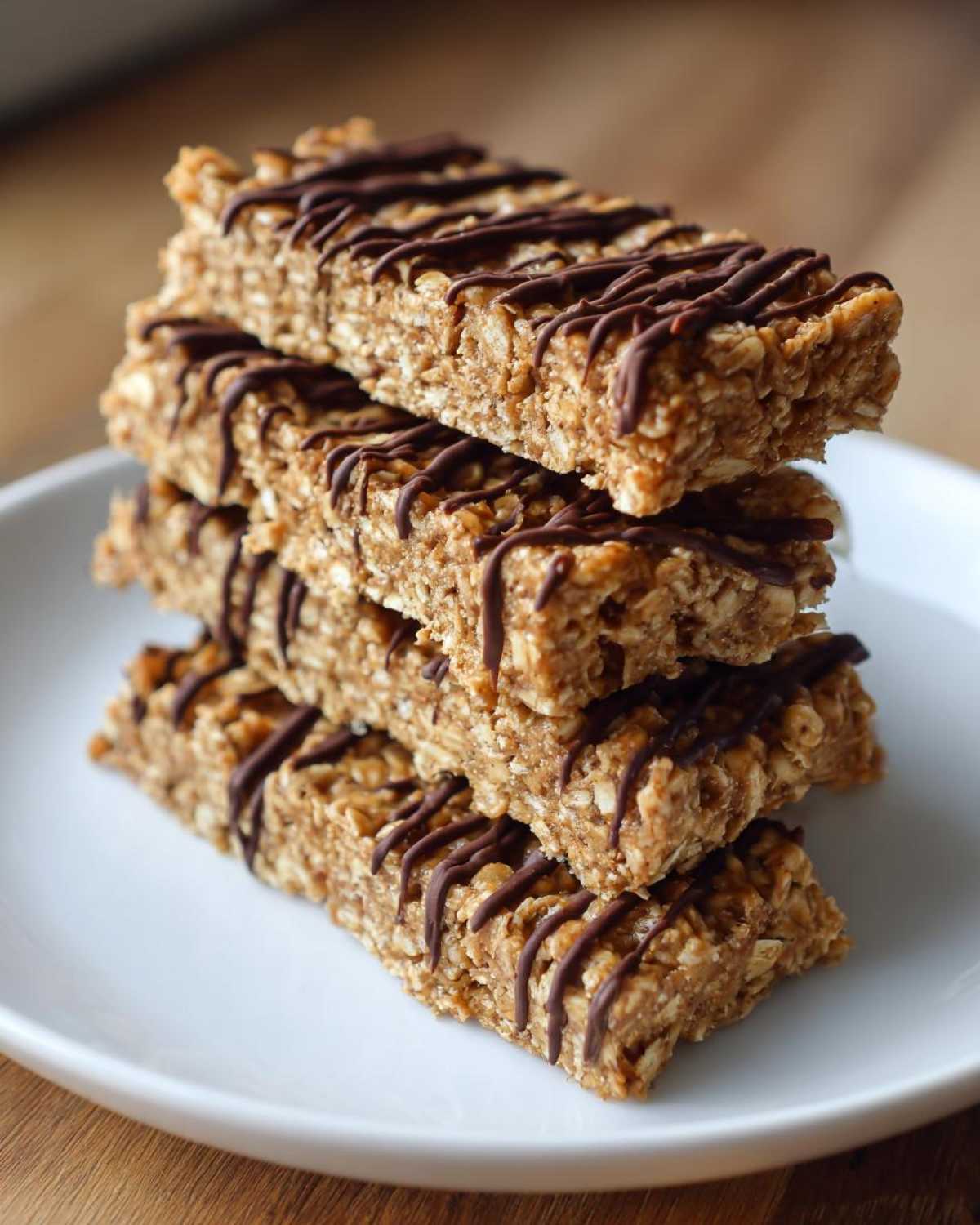 A stack of four homemade protein bars made with oats and drizzled with dark chocolate, resting on a white plate.