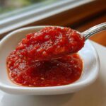 A spoonful of thick, vibrant red homemade pizza sauce being lifted from a small white dipping bowl.