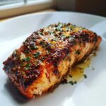 Close-up of a flaky, glazed air fryer salmon fillet topped with garlic and herbs on a white plate.