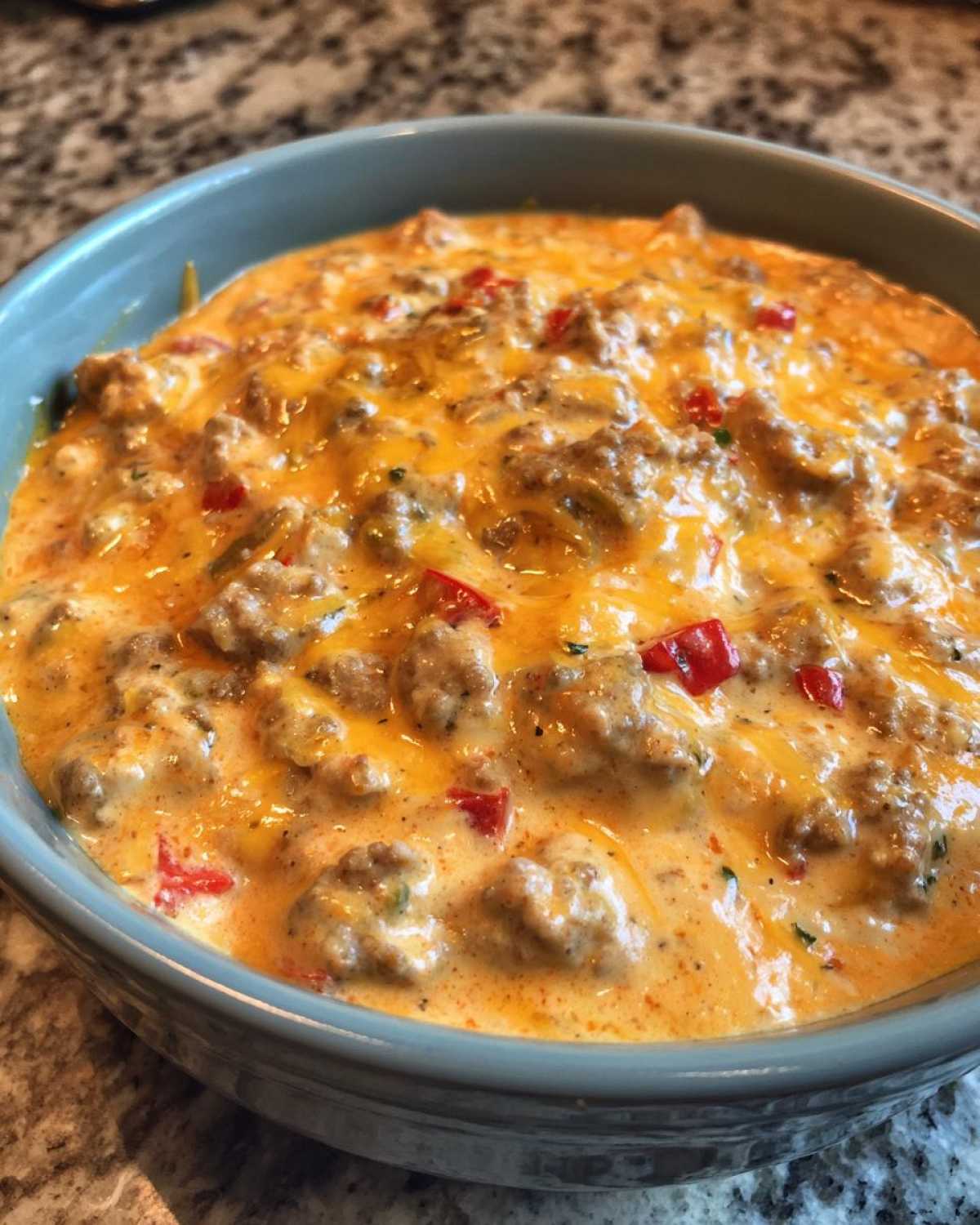 A close-up view of hot, cheesy rotel dip featuring sausage crumbles and diced red peppers in a blue serving bowl.