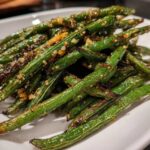 A close-up of perfectly cooked, slightly charred green beans seasoned with minced garlic served on a white plate.