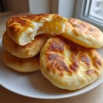 A stack of freshly made, golden-brown pita bread pockets on a white plate, one torn open showing the soft interior.