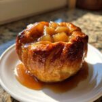 A single, golden-brown baked apple dumpling topped with diced apples and drizzled with caramel sauce.