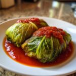 Close-up of two tender cabbage rolls topped with thick tomato sauce, sitting in a pool of sauce on a white plate.