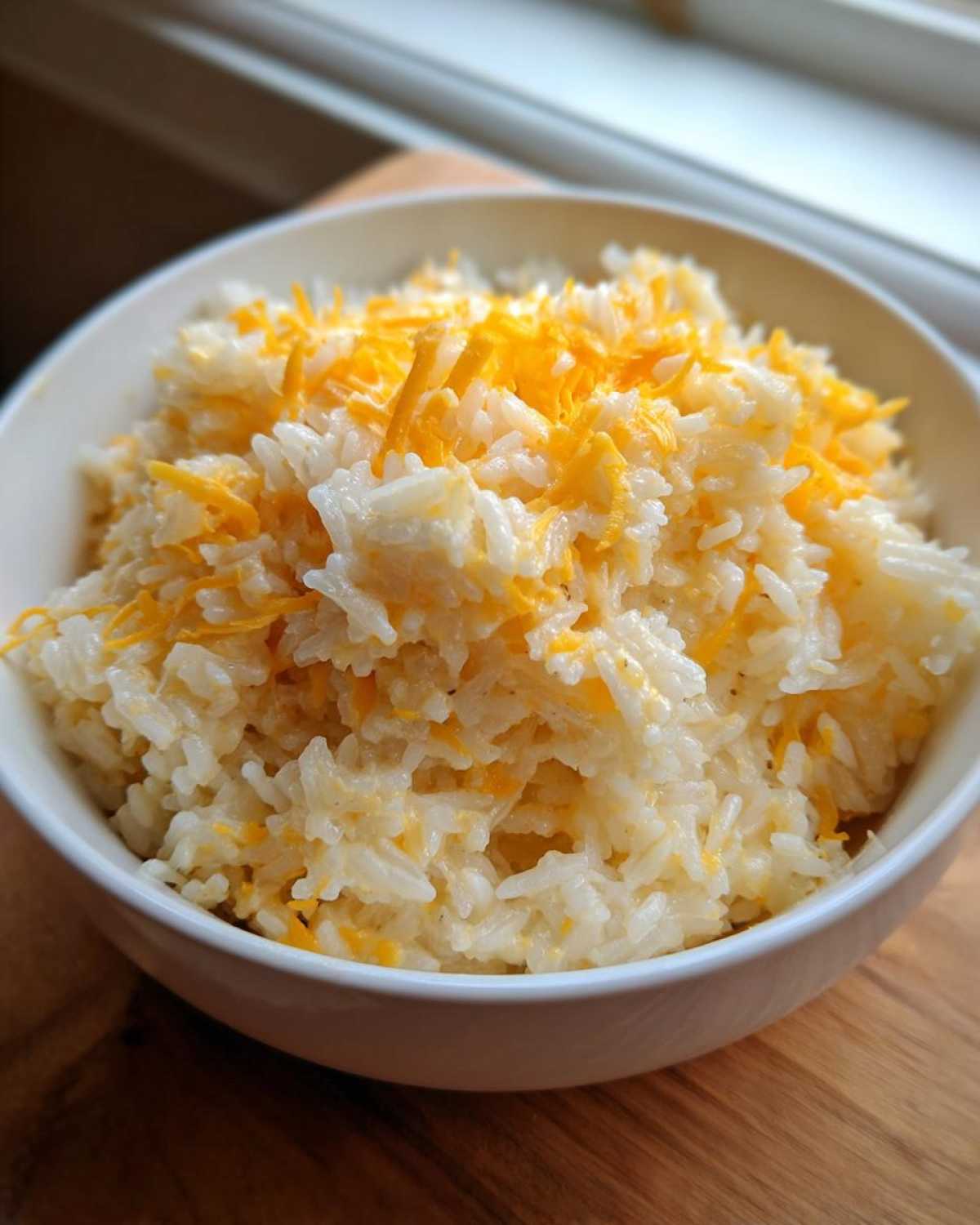 A white bowl filled with fluffy white rice topped with melted and shredded cheddar, showcasing the amazing cheesy rice.