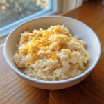 A white bowl filled with creamy, amazing cheesy rice topped with melted shredded cheddar cheese.