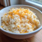 A white bowl filled with fluffy white rice topped generously with shredded cheddar cheese, showcasing the amazing cheesy rice.