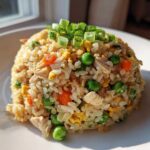 A perfectly molded mound of amazing chicken fried rice featuring visible pieces of chicken, peas, carrots, and egg, topped with green onions.