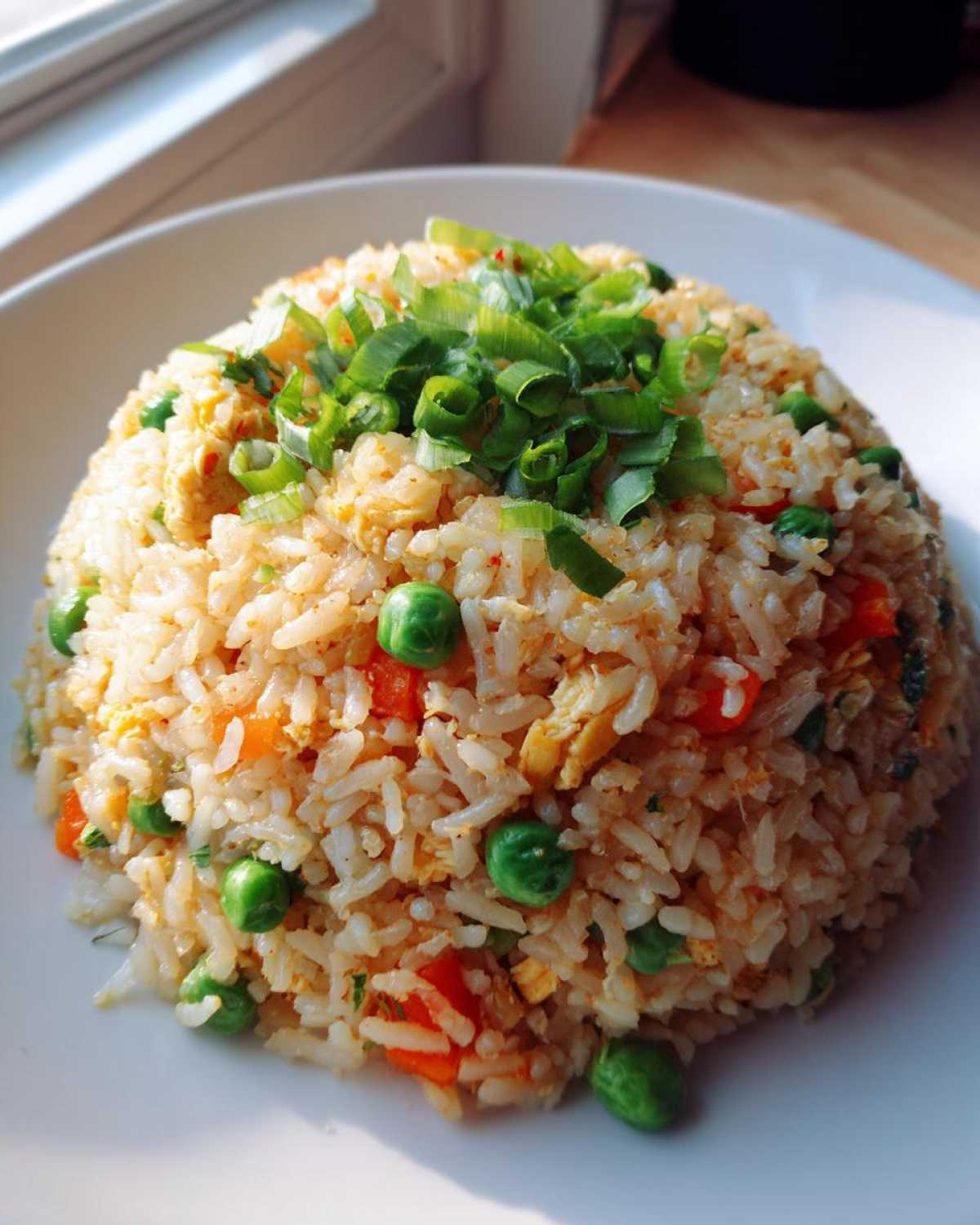 A mound of homemade chicken fried rice mixed with peas and carrots, topped generously with fresh green scallions.