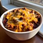 Close-up of a white bowl overflowing with rich chili cheese dip topped with melted cheddar and Monterey Jack cheese.