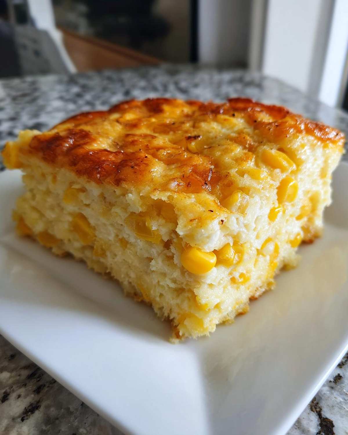 A square slice of moist, baked corn casserole filled with whole corn kernels.