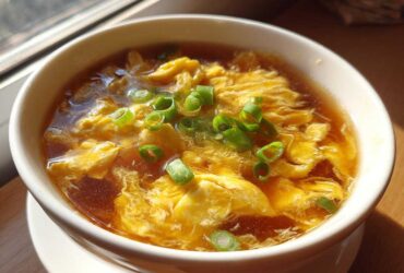 Close-up of a bowl of silky egg drop soup, featuring wispy yellow egg ribbons in a savory broth, topped with fresh green onions.