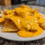 Close-up of tortilla chips smothered in thick, bright orange nacho cheese sauce with visible chili flakes.