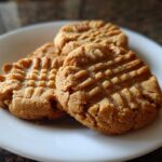 Three golden brown peanut butter cookies with classic crisscross fork marks stacked on a white plate.