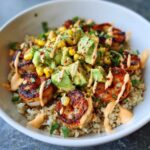 Close-up of a delicious shrimp bowl featuring grilled shrimp, brown rice, avocado chunks, corn, and a creamy orange sauce.