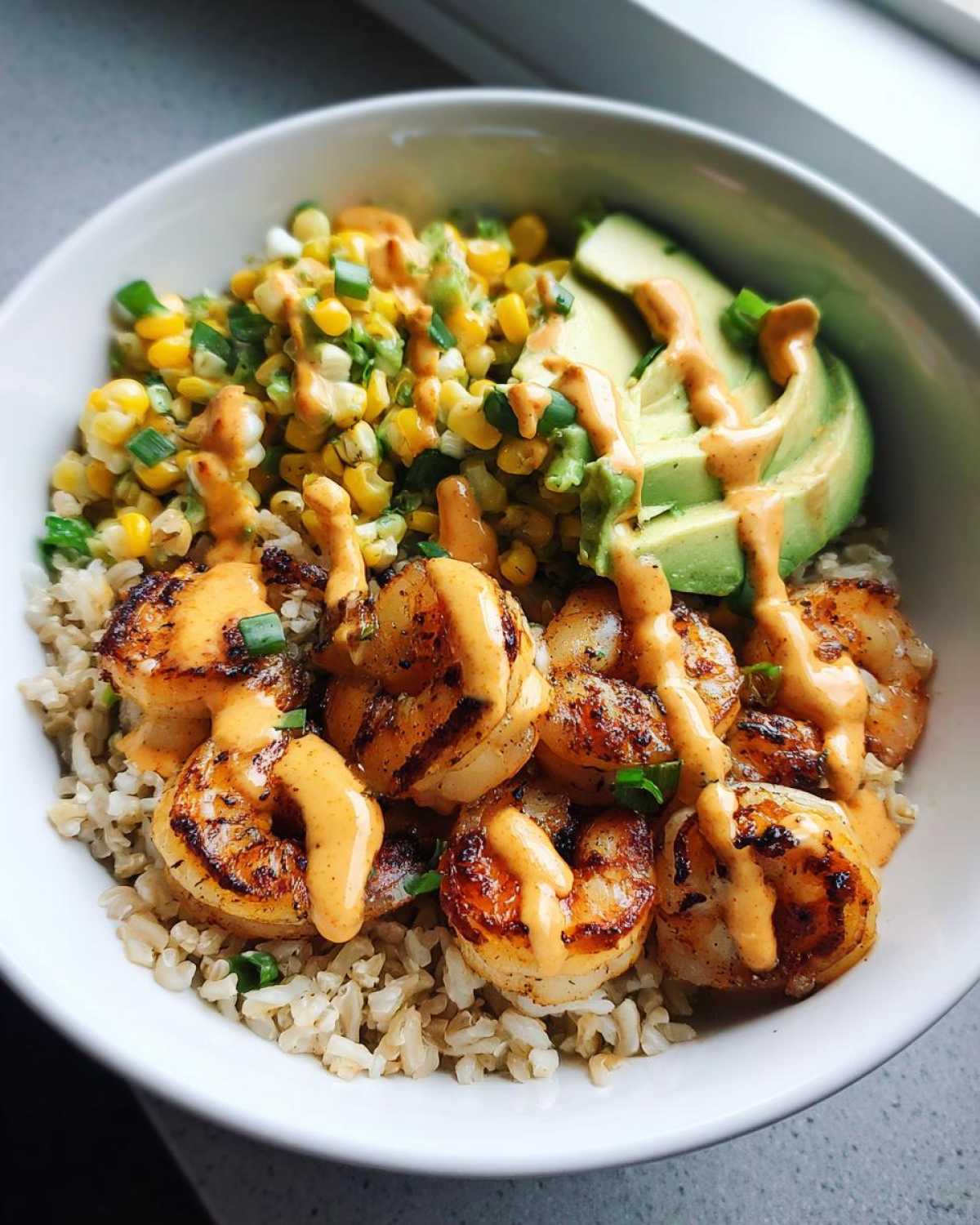 Close-up of a delicious shrimp bowl featuring grilled shrimp, brown rice, corn salsa, and avocado slices drizzled with sauce.