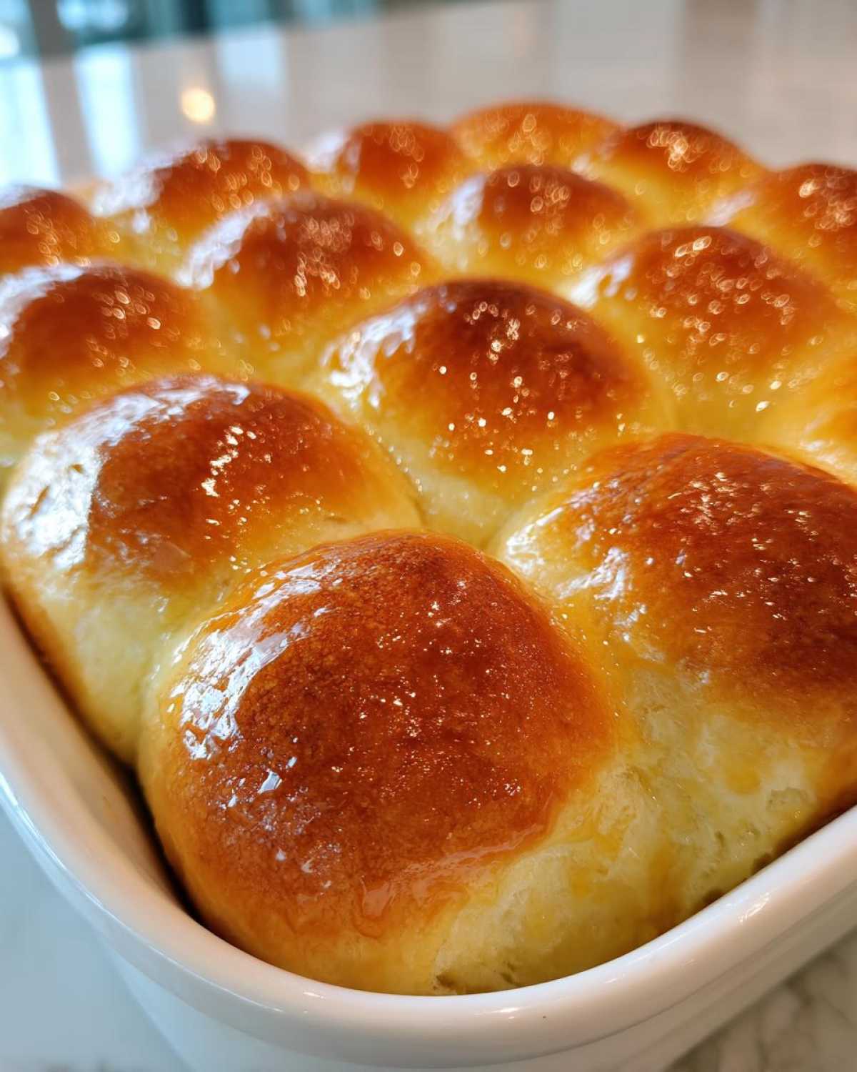 Close-up of a batch of soft dinner rolls baked together in a white dish, featuring a shiny, golden-brown glaze.