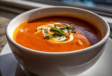 Close-up of a vibrant orange tomato bisque in a white bowl, garnished with a swirl of cream and fresh basil.