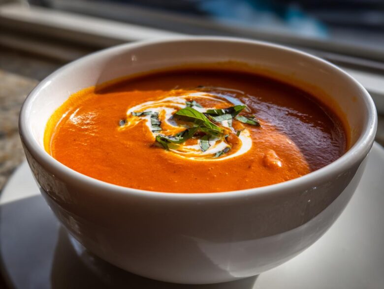 Close-up of a vibrant orange tomato bisque in a white bowl, garnished with a swirl of cream and fresh basil.