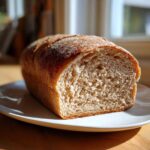 A golden-crusted loaf of homemade whole wheat bread, sliced open to show the soft, airy interior, resting on a white plate.