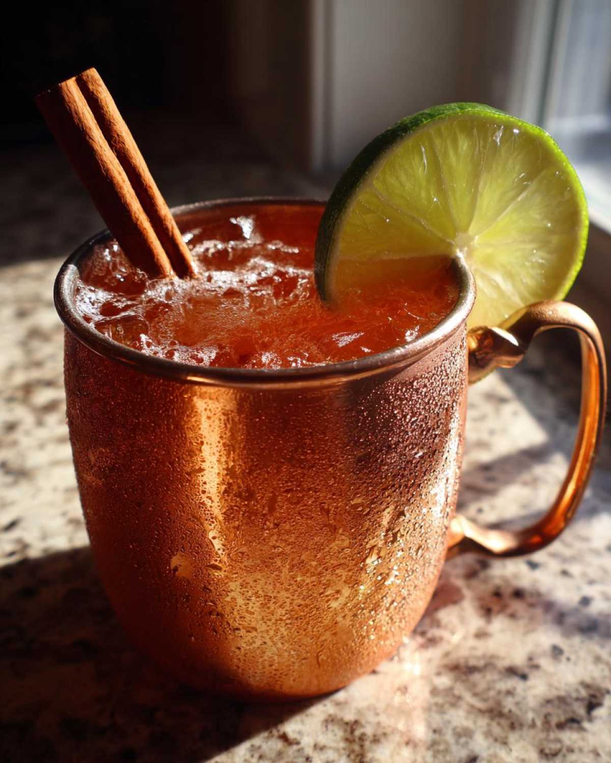 Close-up of a frosty copper mug filled with an apple cider Moscow mule, garnished with lime and cinnamon sticks.