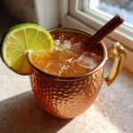 A refreshing apple cider moscow mule served over ice in a hammered copper mug, garnished with a lime wheel and cinnamon stick.