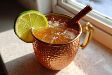 A refreshing apple cider moscow mule served over ice in a hammered copper mug, garnished with a lime wheel and cinnamon stick.