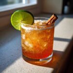 A glass filled with a bubbly, amber-colored apple cider moscow mule, garnished with a lime wheel and cinnamon stick.