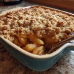 Close-up of a golden brown apple crisp recipe baked in a teal ceramic dish with a spoon digging in.
