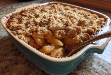 Close-up of a golden brown apple crisp recipe baked in a teal ceramic dish with a spoon digging in.