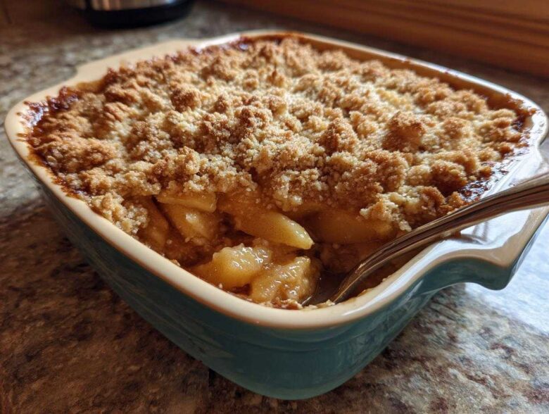 Close-up of a golden brown apple crisp recipe baked in a teal ceramic dish with a spoon digging in.