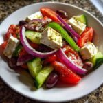 A vibrant, close-up serving of authentic greek salad featuring large chunks of feta, tomatoes, cucumbers, red onion, and Kalamata olives.