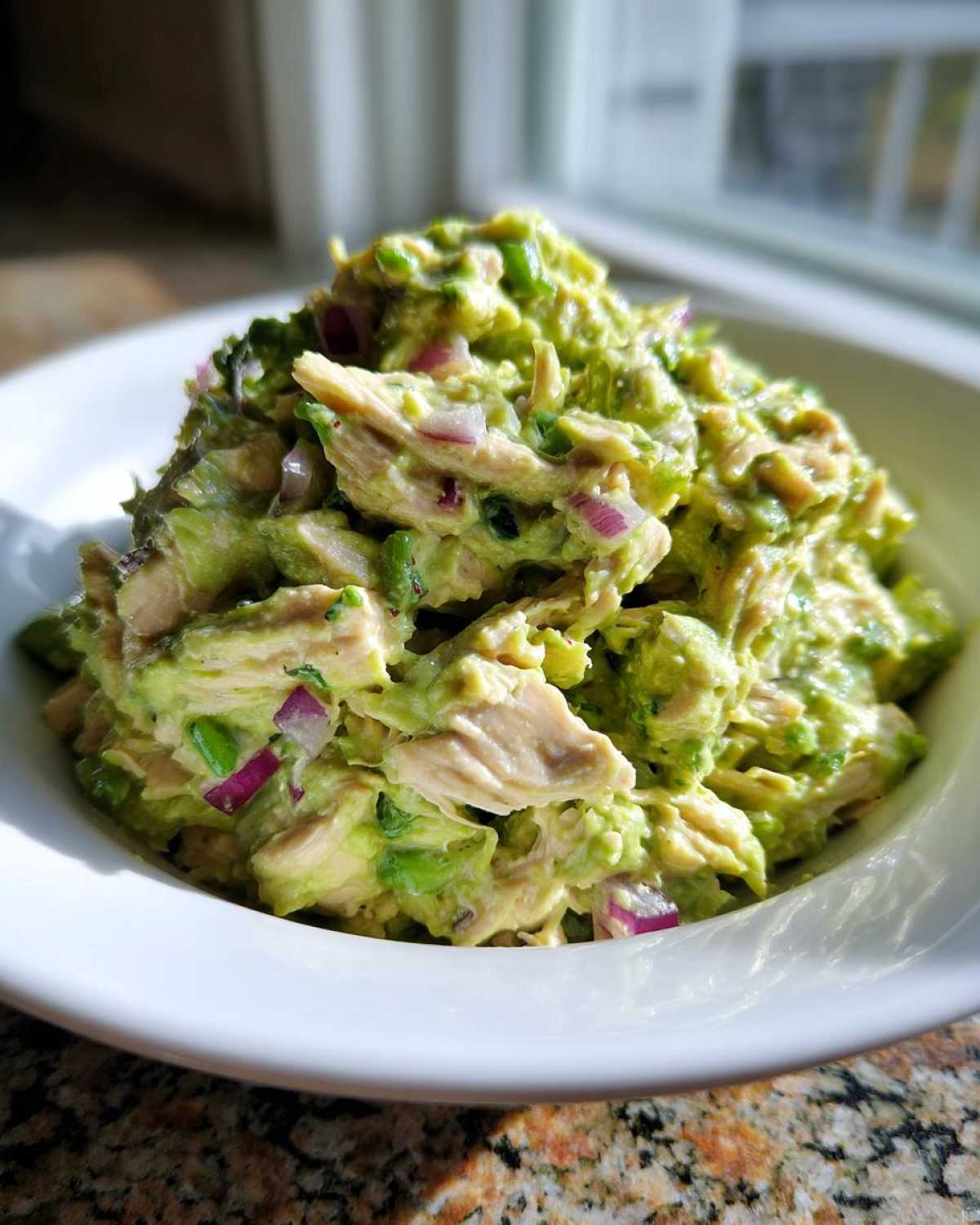 Close-up of creamy avocado chicken salad mixed with chunks of chicken and red onion in a white bowl.