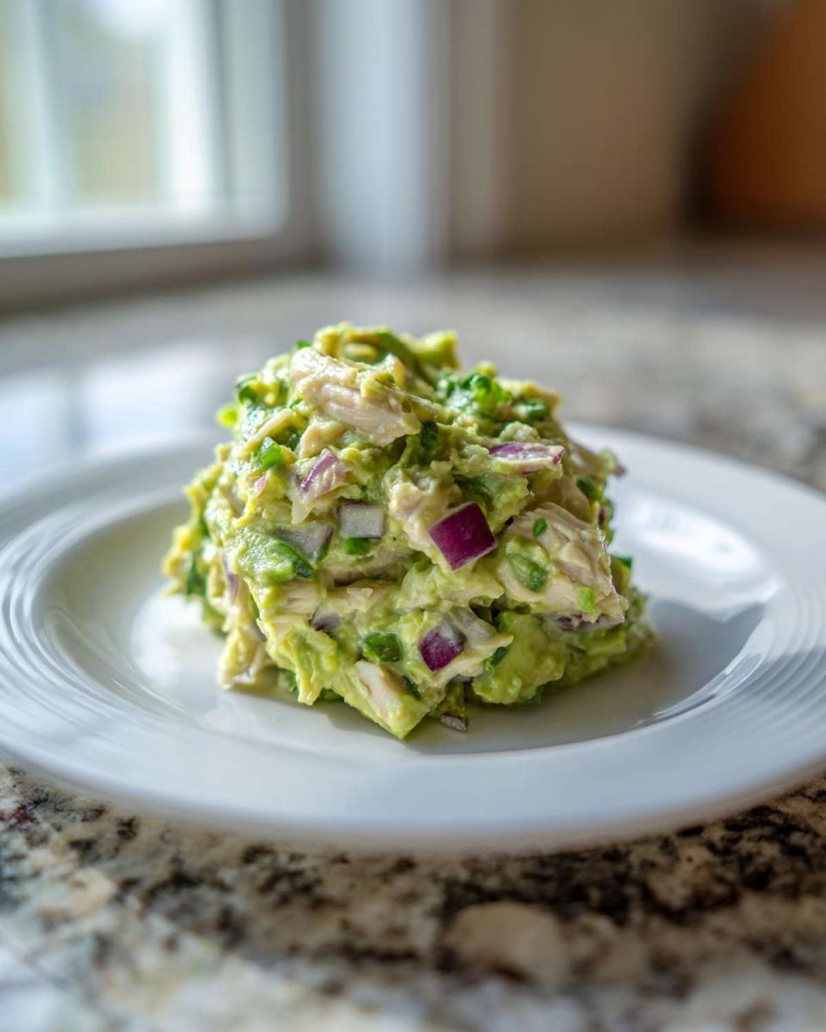 A scoop of creamy avocado chicken salad mixed with chunks of red onion served on a white plate.