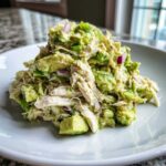 A mound of creamy avocado chicken salad mixed with chunks of avocado and red onion on a white plate.