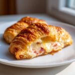 Close-up of a baked ham and cheese croissant, split open to show melted, gooey cheese and ham filling.