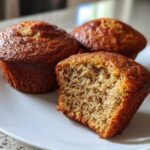 Three golden-brown banana protein muffins on a white plate, one is broken open showing the moist crumb.