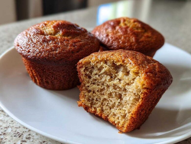 Three golden-brown banana protein muffins on a white plate, one is broken open showing the moist crumb.