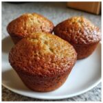 Close-up of three freshly baked banana protein muffins with golden brown, slightly glossy tops resting on a white plate.
